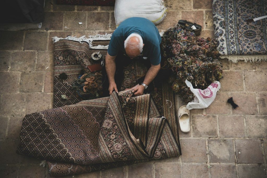 Man folding a patterned rug on a tiled floor.