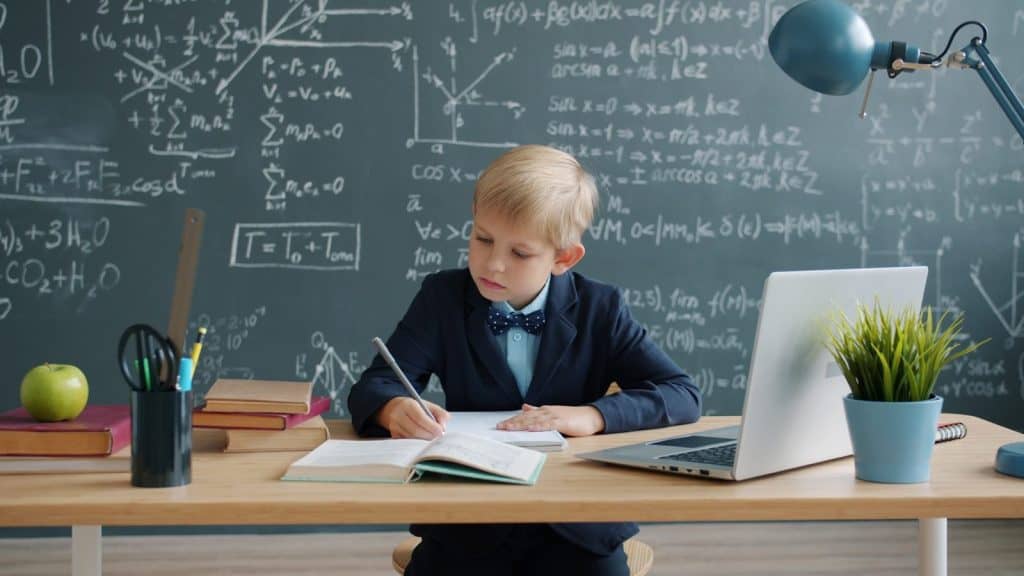 Young boy in suit writing at desk with chalkboard.